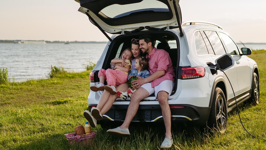 A family with two daughters sitting in the boot of hatchback parked by a lake