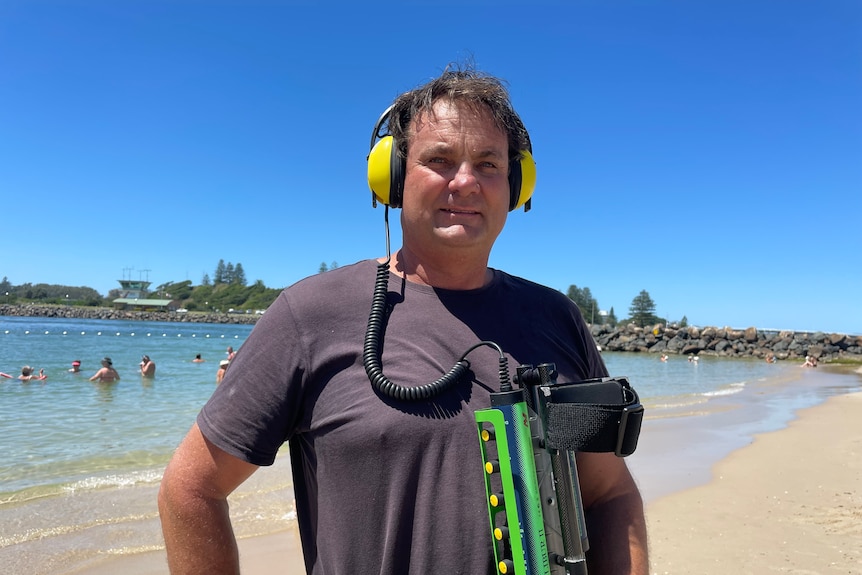 A man wearing a t-shirt stands at the beach wearing headphones and holding a metal detector.