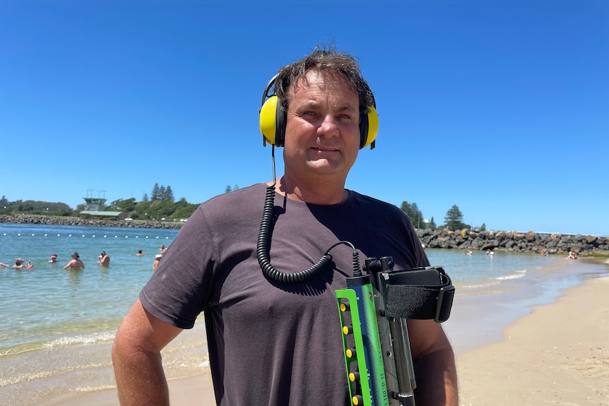 Un homme portant un t-shirt se tient à la plage avec des écouteurs et tenant un détecteur de métaux.