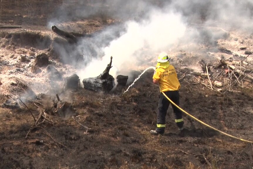 Man in yellow fire gear stands on burnt ground, directing a hose