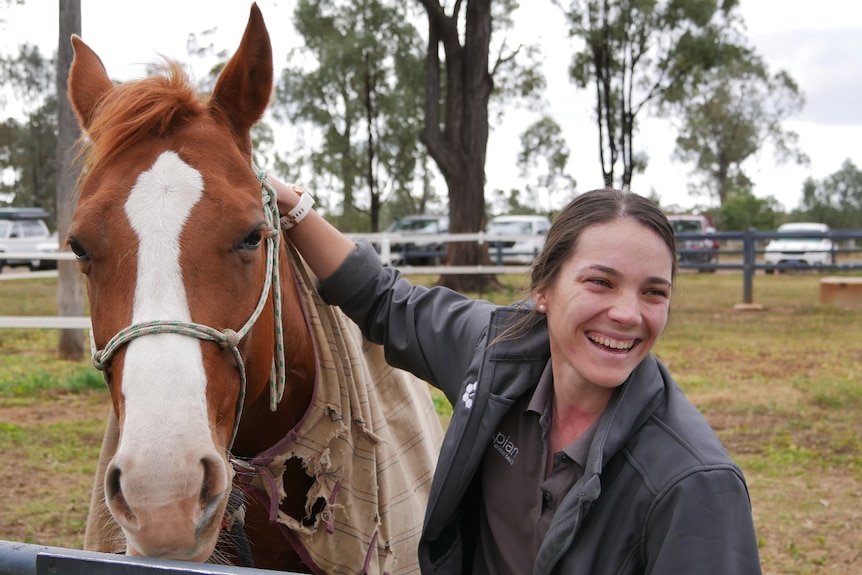 Woman smiling, hand on brown and white horse, trees behind.