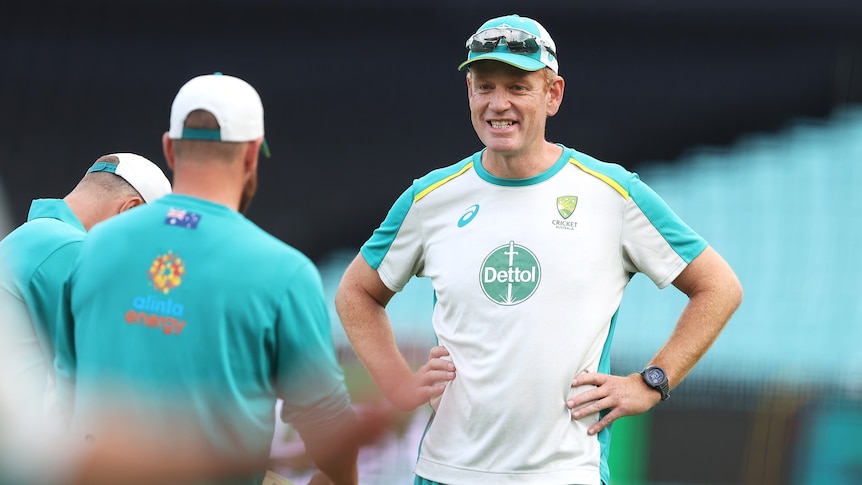An Australian cricket coach stands smiling with hands on hips talking to a player before a game. 