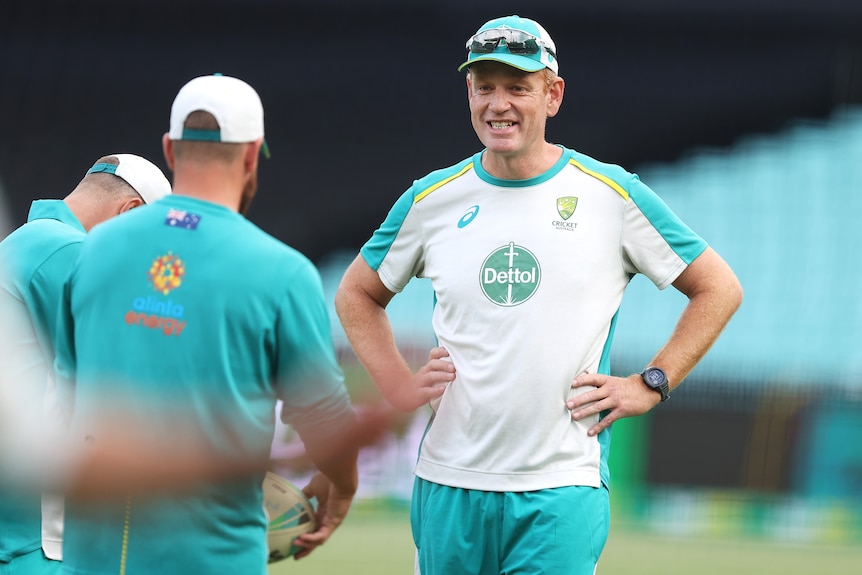 An Australian cricket coach stands smiling with hands on hips talking to a player before a game. 