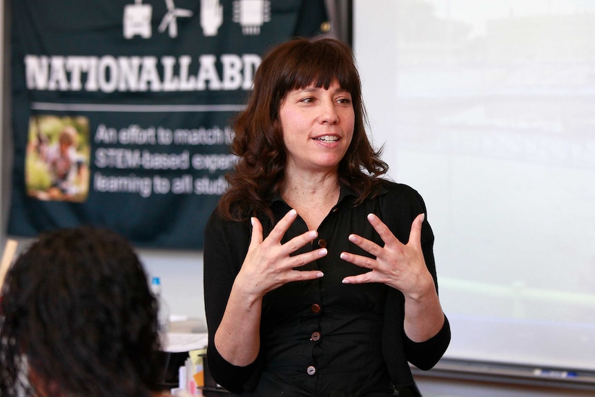 A woman with dark hair gestures with her hands as she speaks.