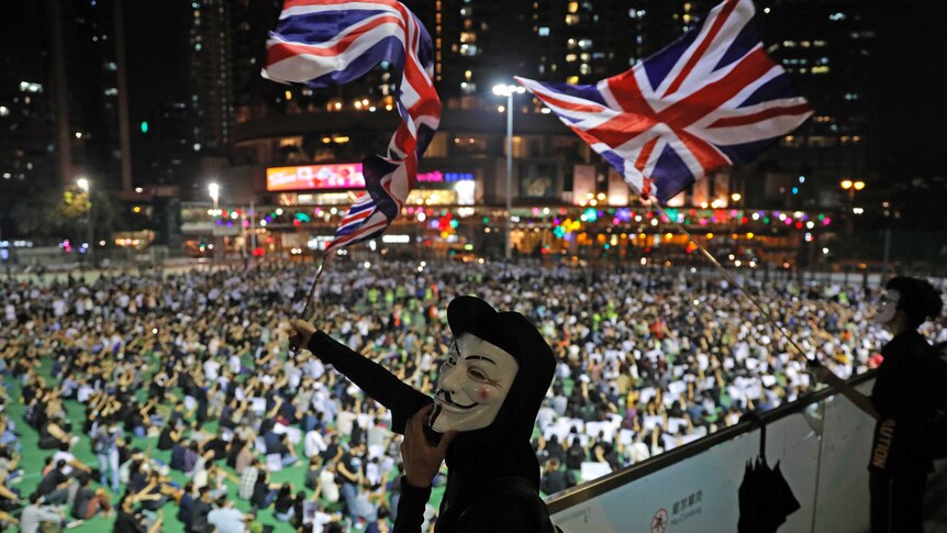 Anti-government protesters carry British flags as a man wears a white mask in front of the crowd.