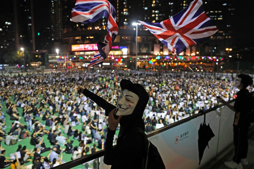 Anti-government protesters carry British flags as a man wears a white mask in front of the crowd.