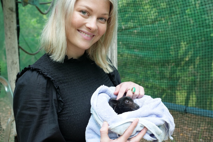 A blonde woman smiling at the camera holding a small black bat in a towel