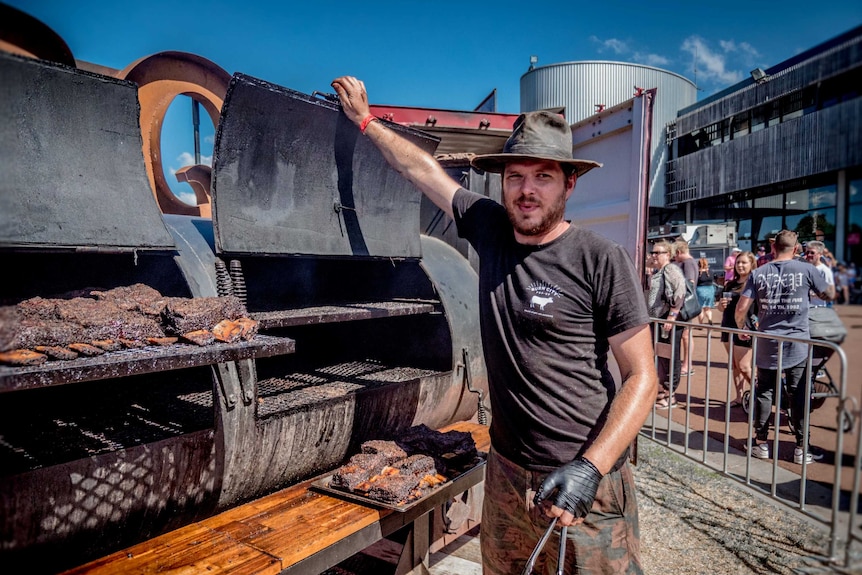 A bearded man in a cattle hat smiles while holding open a large smoker barbecue at the 2019 Meatstock event in Melbourne.