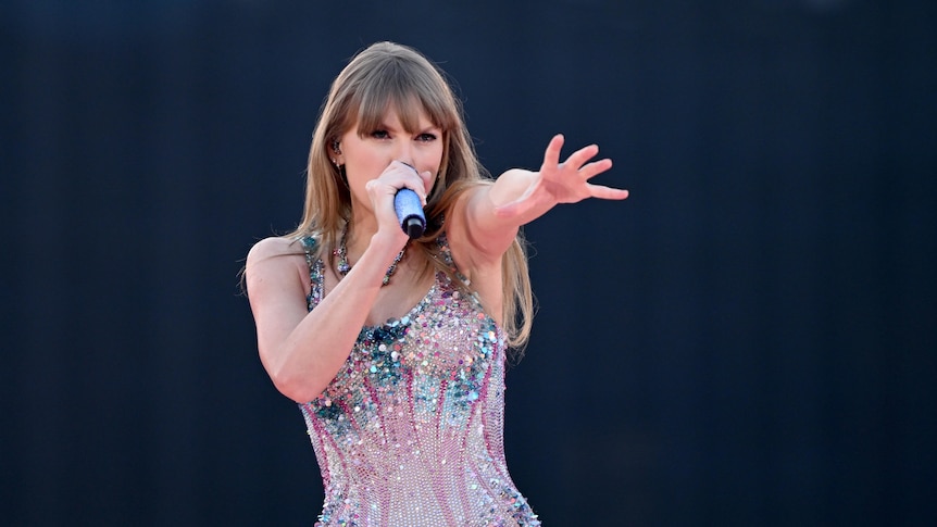 Taylor Swift holds a microphone and dances in a glittering bodysuit.
