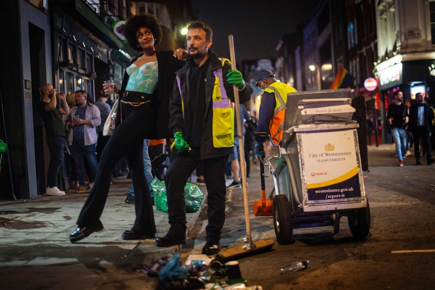 A woman holding a drink poses for a photograph with a binman who is holding a broom.