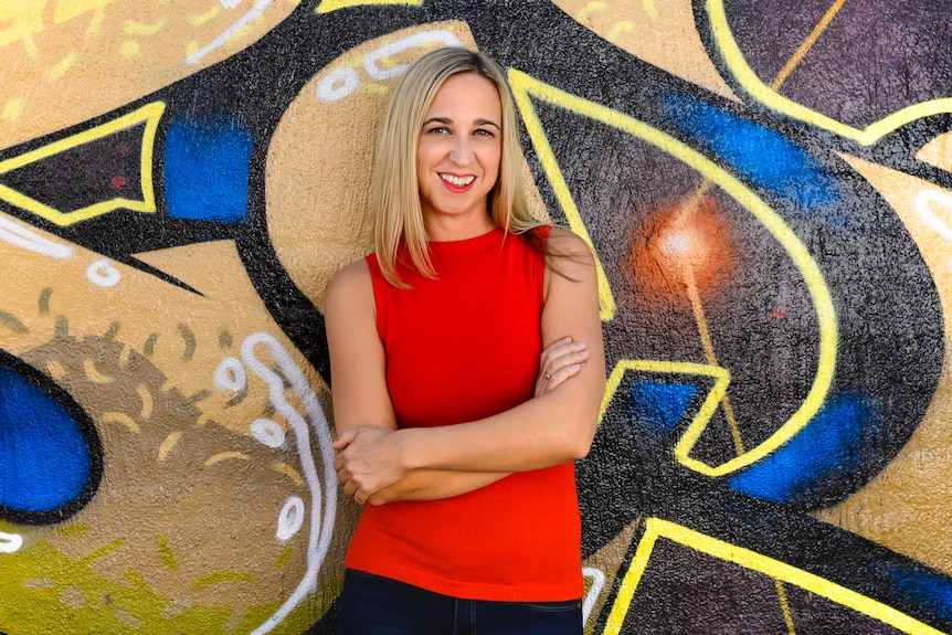 A blonde woman wearing a red top and jeans stands in front of a colourful wall, smiling.