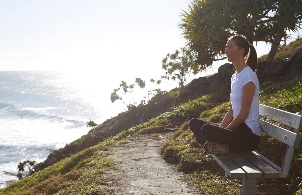 Woman sits on a bench by the coast cross legged and looks out to sea.