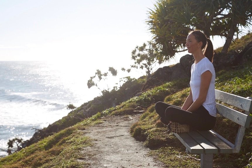 Woman sits cross legged on a bench by the coast and looks out to sea.