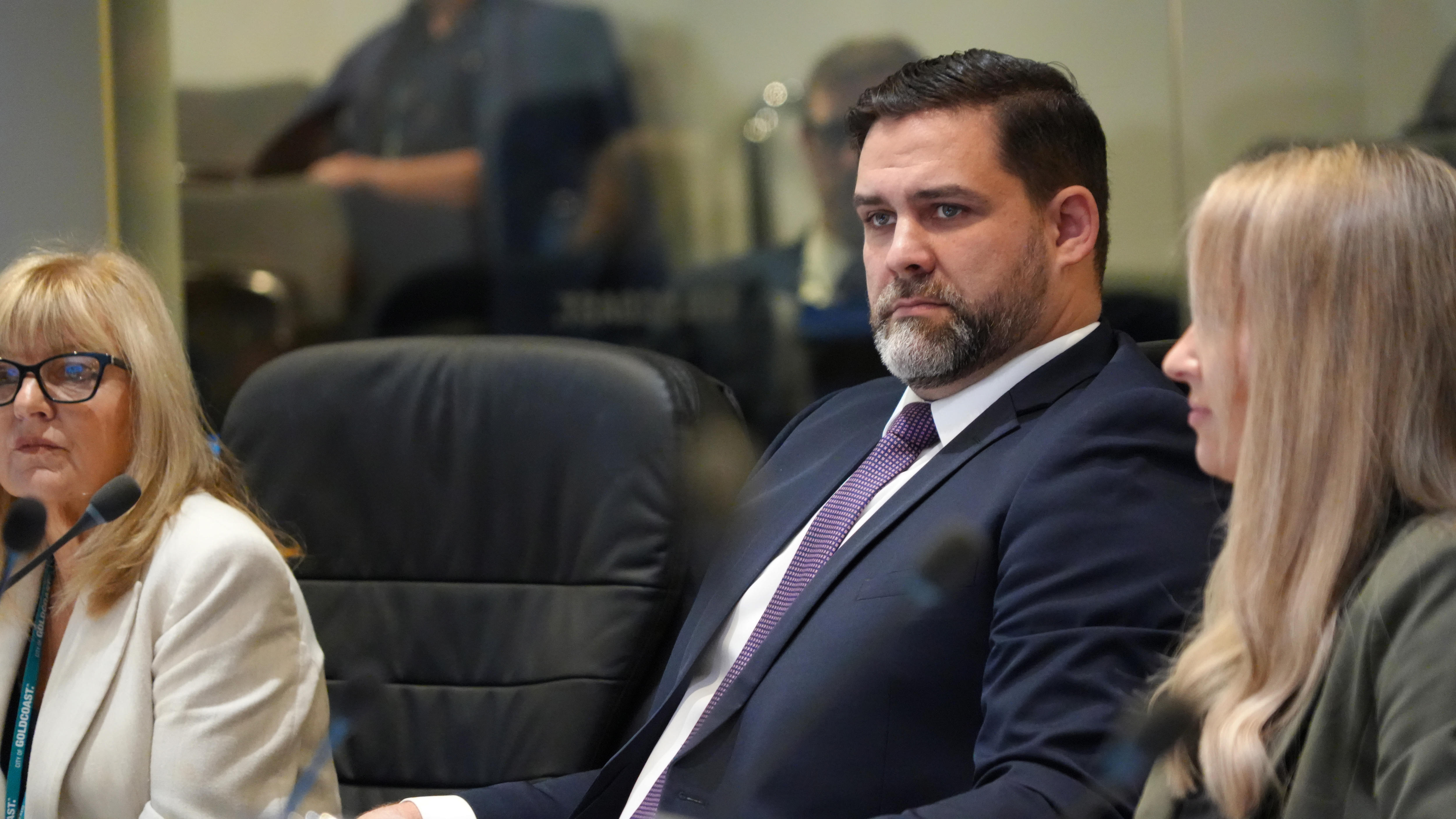 A dark-haired, bearded man in a dark suit sits between two blonde women in a council chamber.