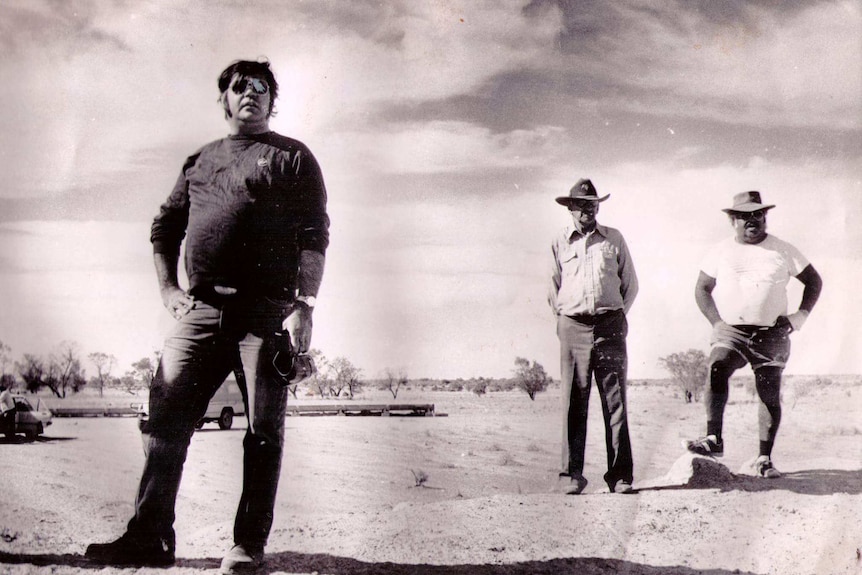 Black and white photog of a tall aboriginal man in in sunglasses the foreground, two aboriginal men in Akubra hats in background