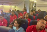 Tongan men in red shirts and a Japanese woman sit in an airline.