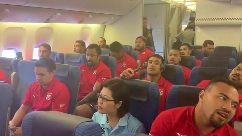 Tongan men in red shirts and a Japanese woman sit in an airline.