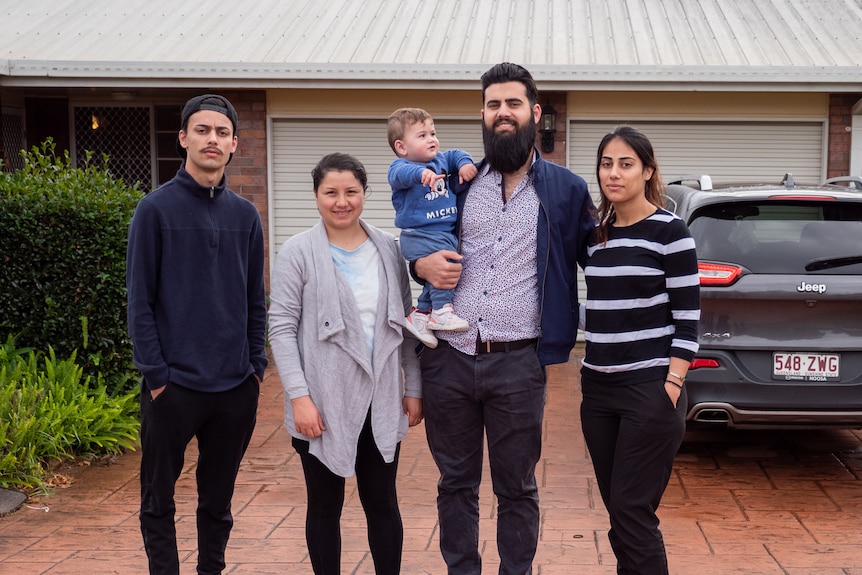 A dark-haired, bearded man and his family stand out the front of their house.