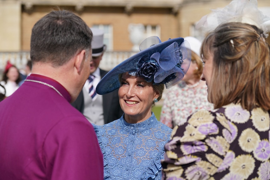 A woman with a blue dress and fascinator is pictured smiling as she speaks to people whose backs are turned to the camera.
