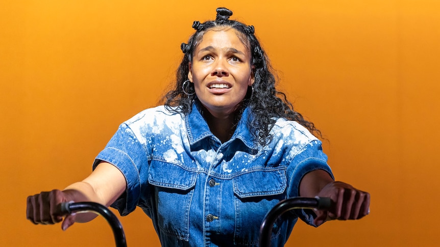 On stage, Zahra Newman, a Jamaican Australian actor, is perched on a bike, with a concerned expression.