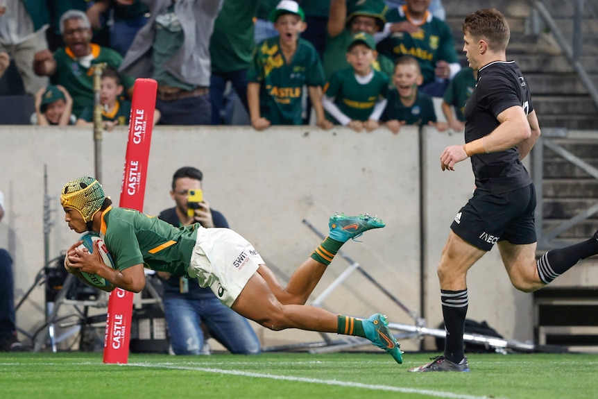 A South African rugby union player dives through the air to score near the corner post as a New Zealand player trails behind. 