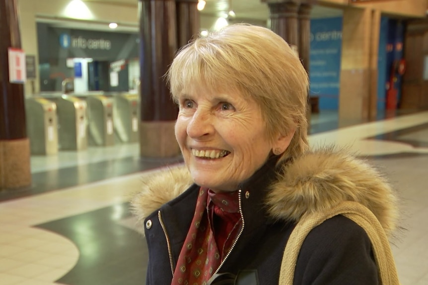 A smiling woman with short blonde hair and a thick jacket standing in a railway station