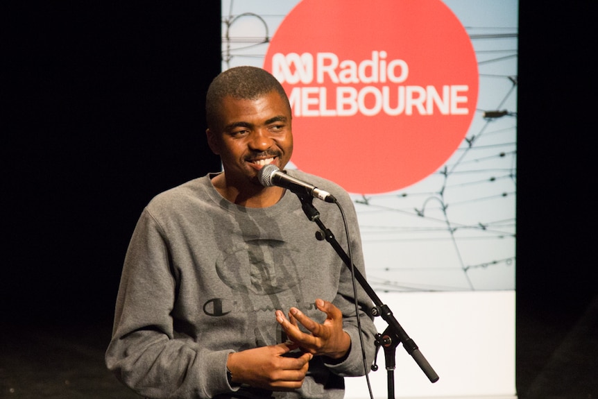 A man wearing a grey sweater smiles as he talks into a microphone. Banner in background for ABC Radio Melbourne.