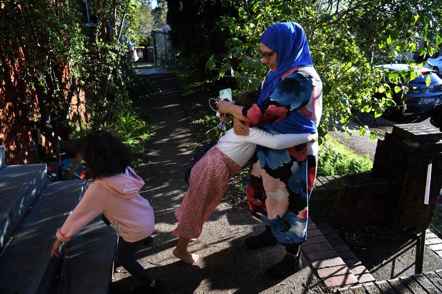 A woman wearing a bright long-sleeve dress and neon blue headscarf as well as glasses hugs a young child in a home frontfootpath
