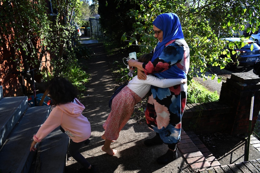 A woman wearing a bright long-sleeve dress and neon blue headscarf as well as glasses hugs a young child in a home frontfootpath