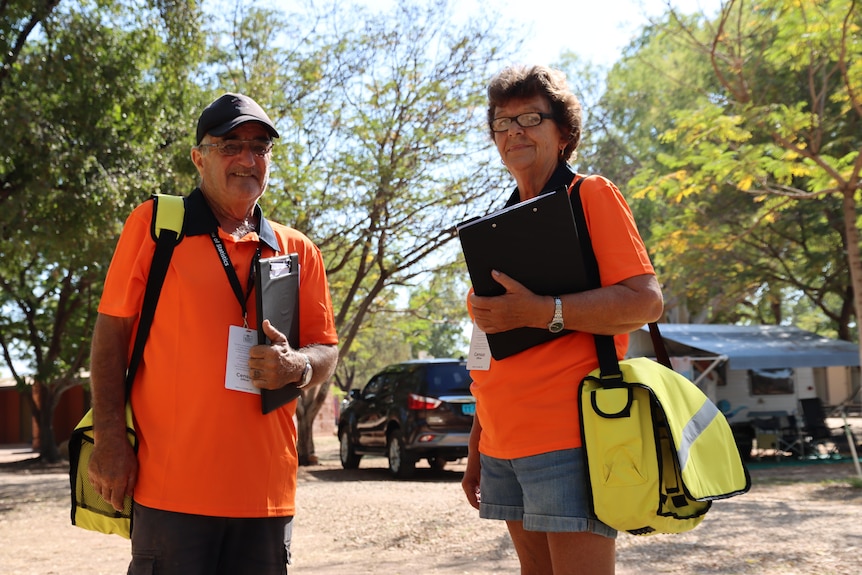 Barry and Liz Price stand in bright orange Census shirts holding clipboards in a caravan park. 