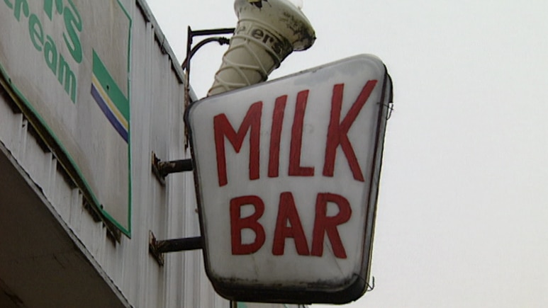 A slightly weathered white 'Milk Bar' sign with red lettering and broken ice cream cone outside shopfront.