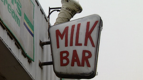 A slightly weathered white 'Milk Bar' sign with red lettering and broken ice cream cone outside shopfront.