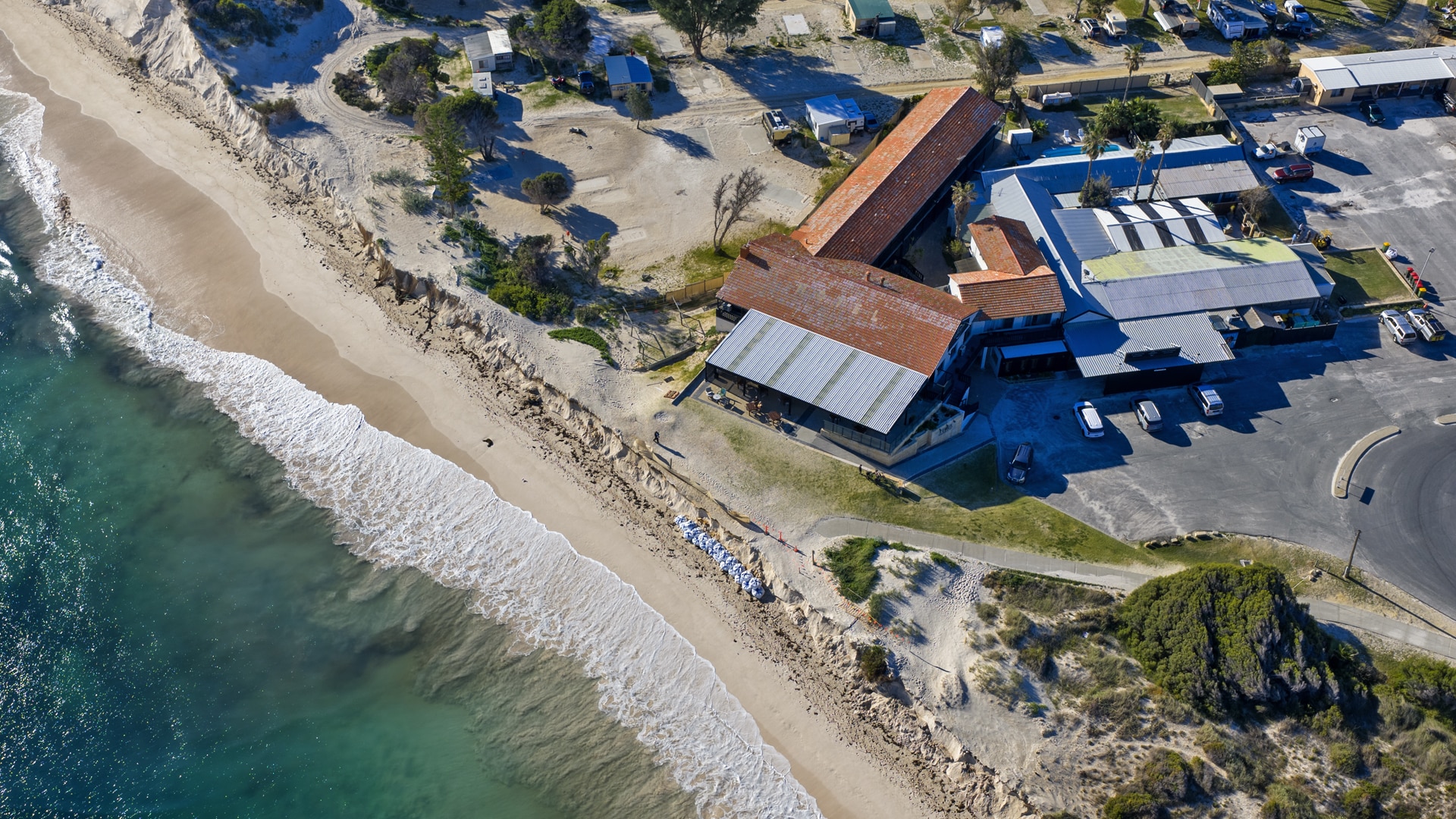 A drone view of ocean at an eroded beach, with a building sitting about 5 metres from a sand cliff.
