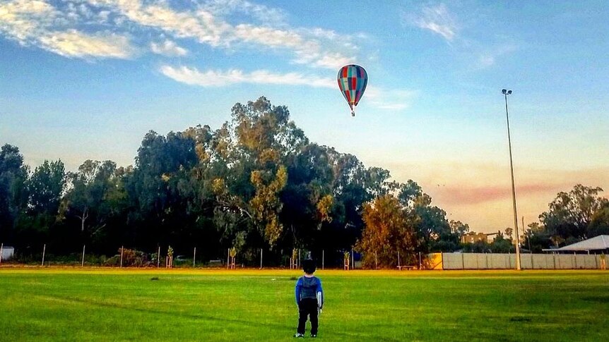 View from behind of a boy standing in a park watching a hot air balloon in the distance.