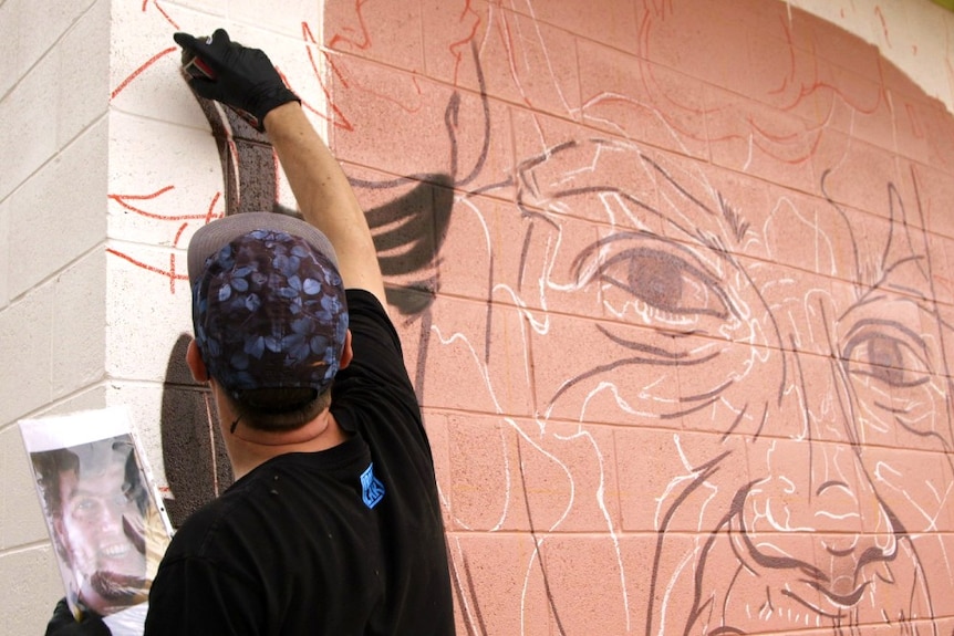 A man paints a mural on a wall. He has his back to the camera