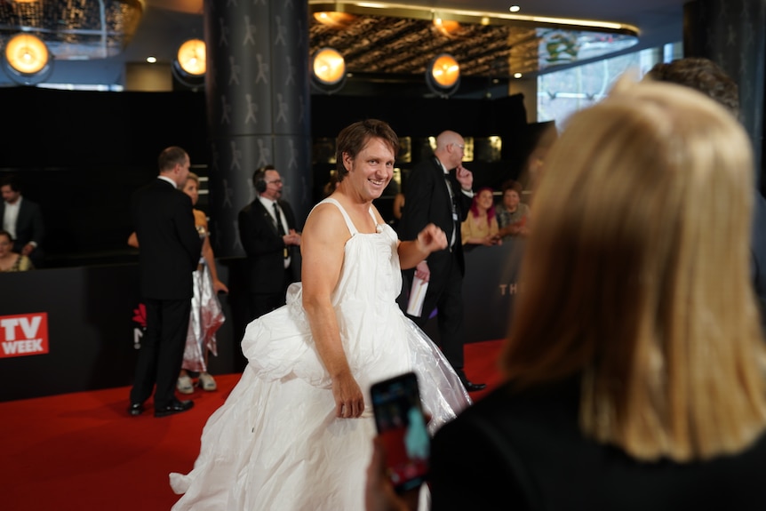 A man stands on a red carpet wearing a white dress.