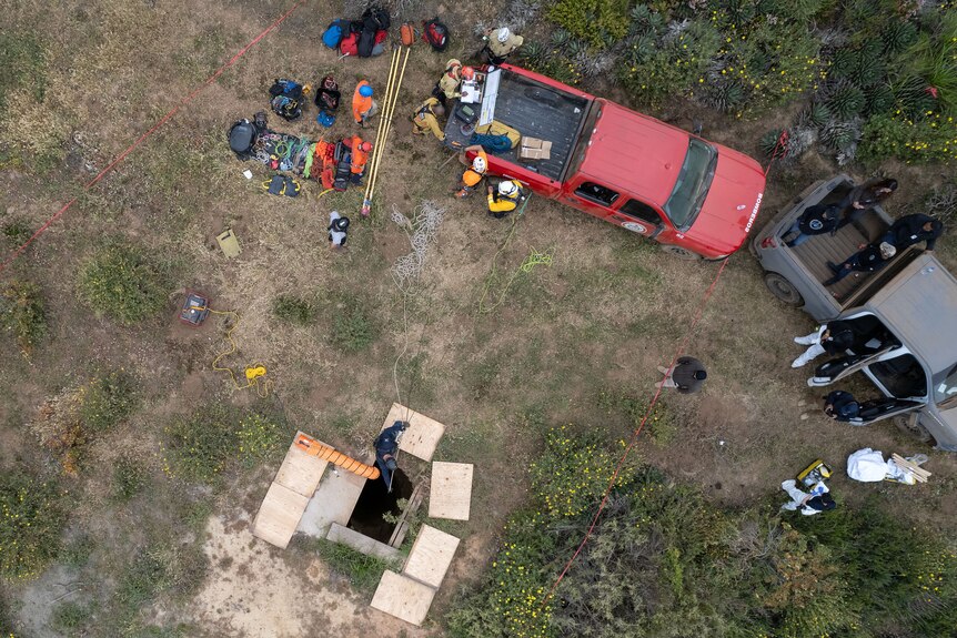 Aerial shot of a person climbing into a hole in the ground surrounded by utes and other people. 