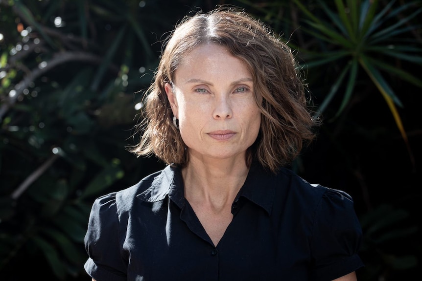 A stern-faced woman with shoulder-length brown hair, wearing a black shirt