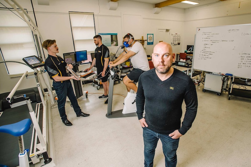 Dean Mills looks stern while a volunteer's lungs are tested on an exercise bike behind him.