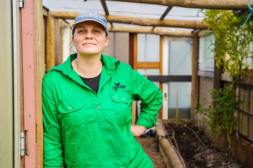 A woman in a green shirt and cap stands in the doorway of a greenhouse smiling.