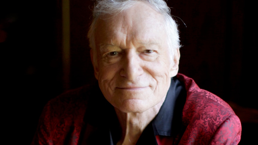 Playboy magazine founder Hugh Hefner smiles at the camera while wearing a red bathrobe.