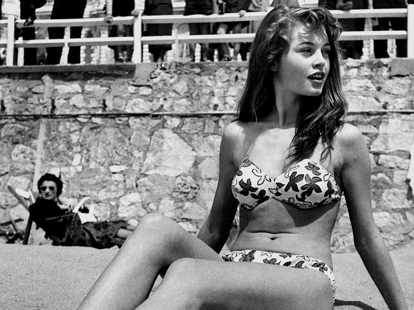A black and white image shows a woman sitting upright on a beach in a flower-patterned white bikini as a crowd watches on.