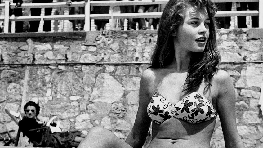 A black and white image shows a woman sitting upright on a beach in a flower-patterned white bikini as a crowd watches on.