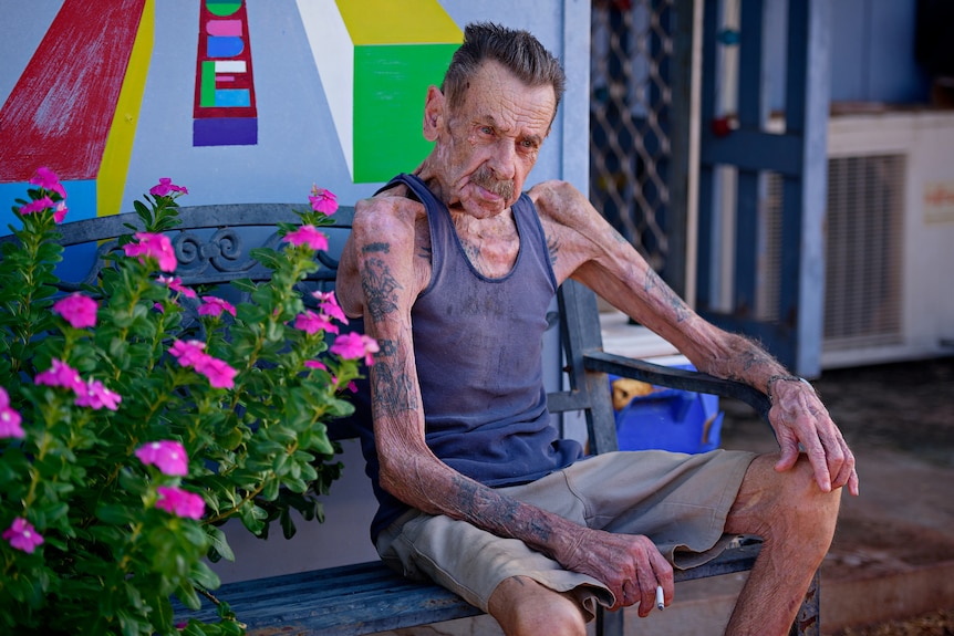 An elderly, very thin man in a singlet top and shorts, sitting down outside a building.