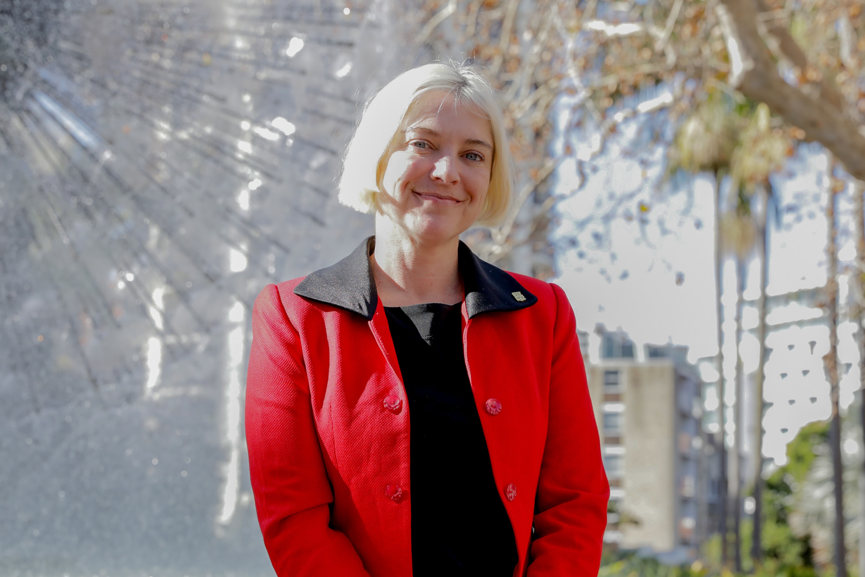 A blonde white woman wearing a red jacket smiles directly at the camera in front of a water fountain.