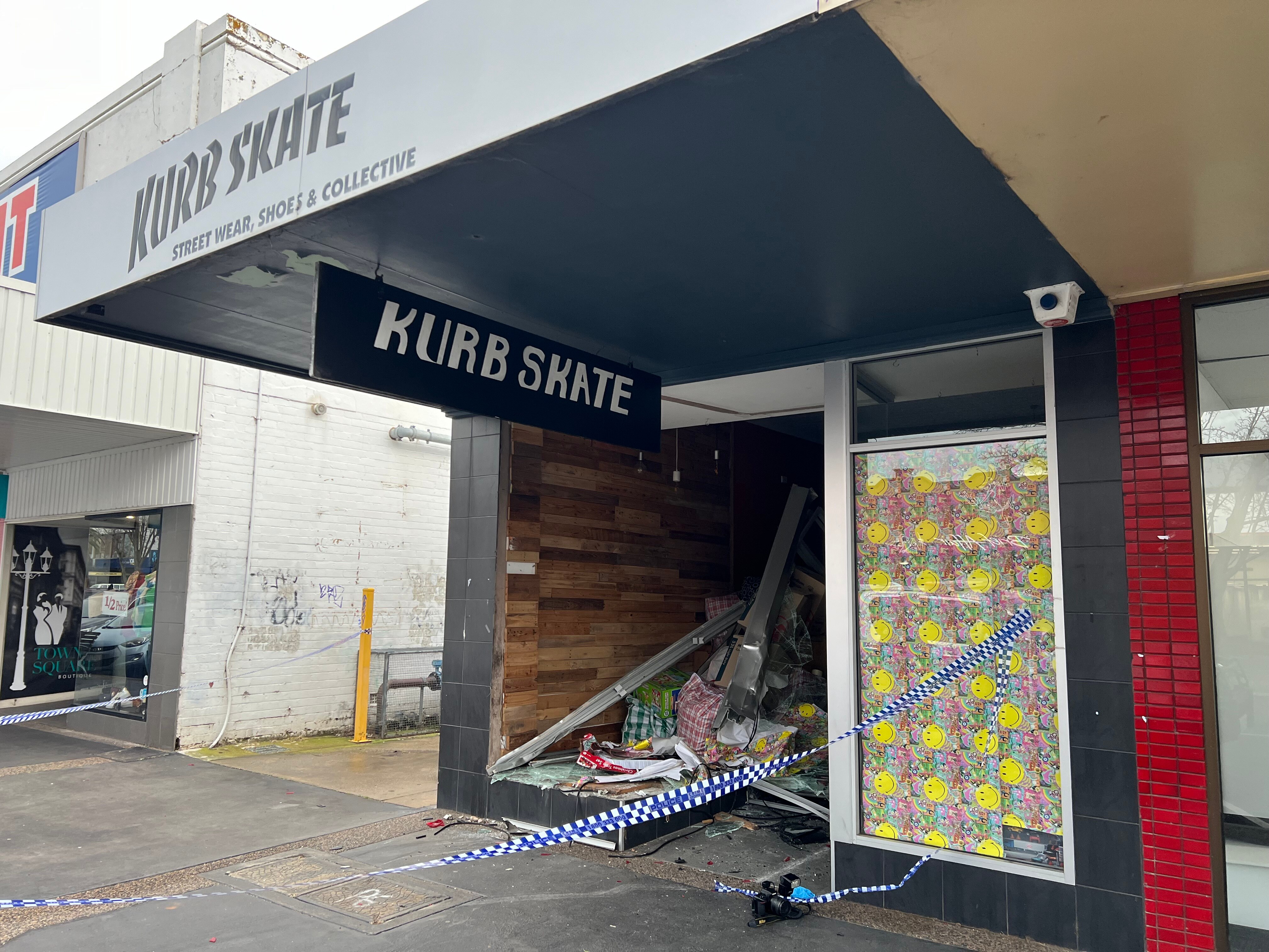 Smashed main window of store with police tape outside and broken glass on pavement.