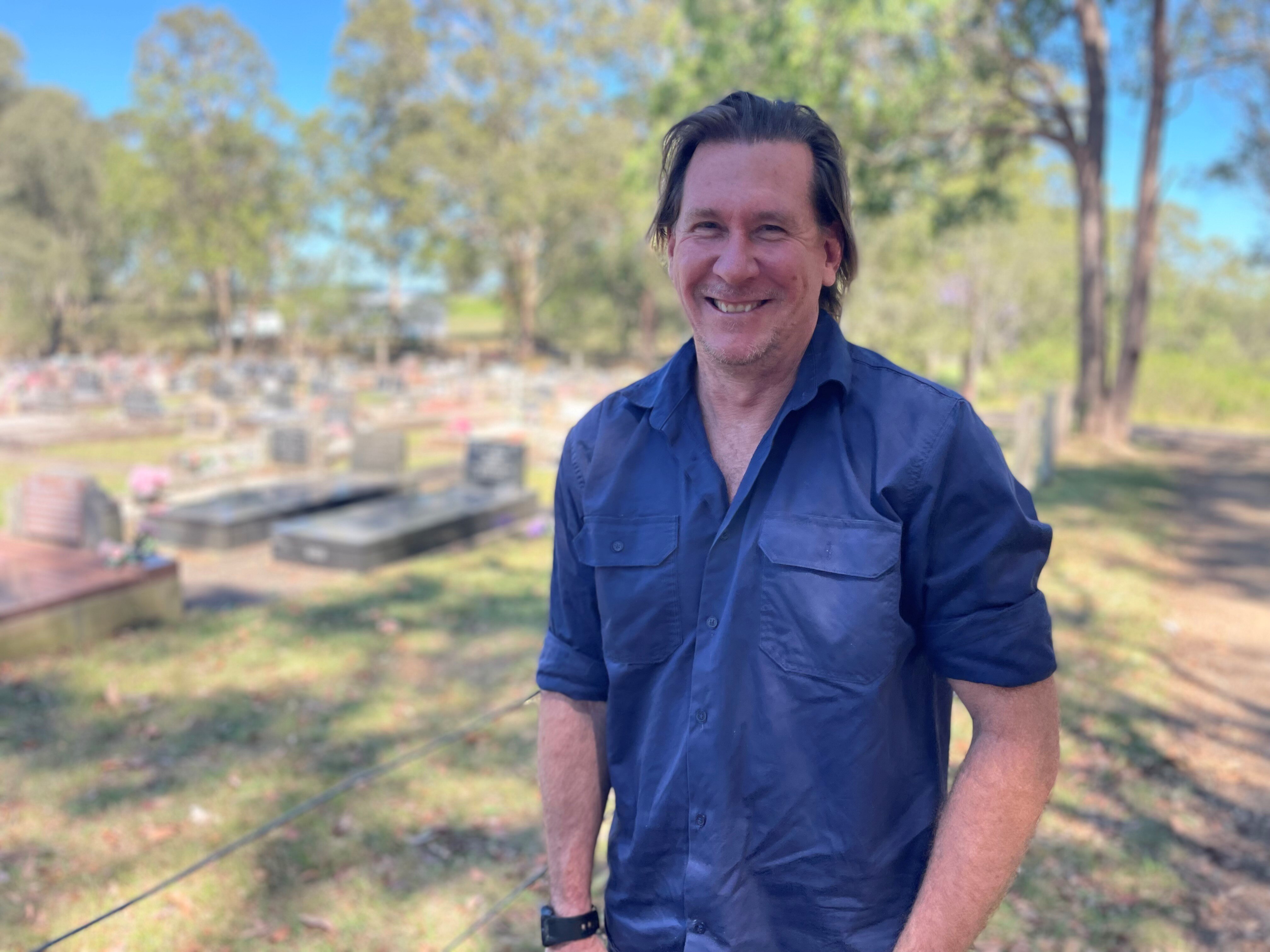 A man in a dark blue shirt, standing on the edge of a cemetery with bushland in the background.