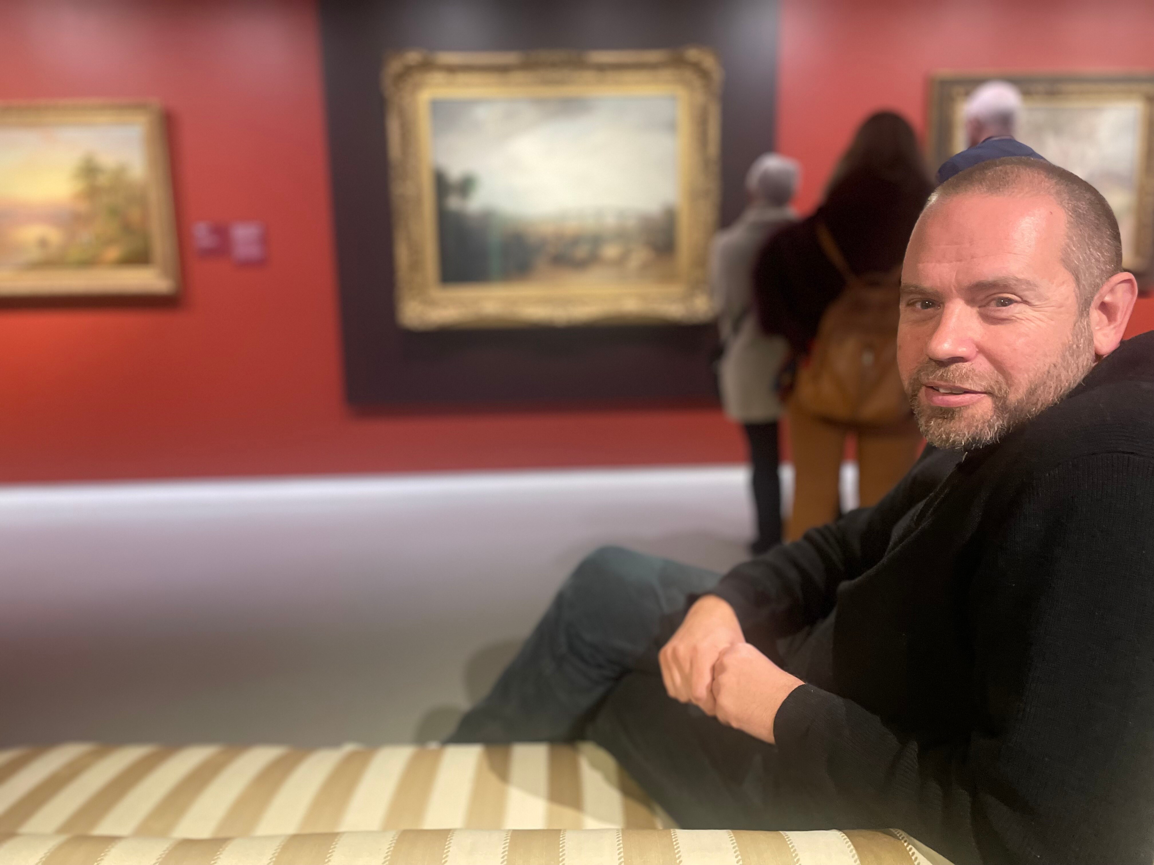 man with moustache and beard wearing black sitting on chaise lounge at the Turner exhibition  