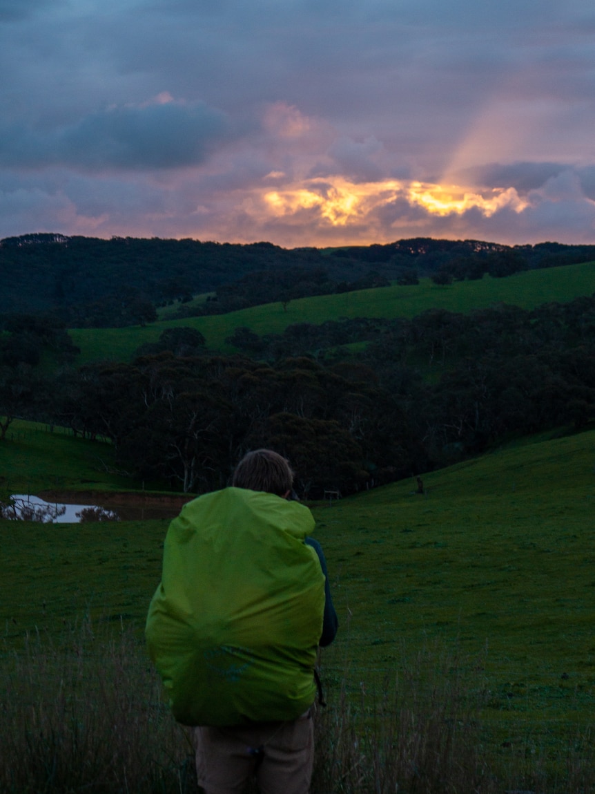 A boy's back with a large green camping bag in front of hills and a colorful sky