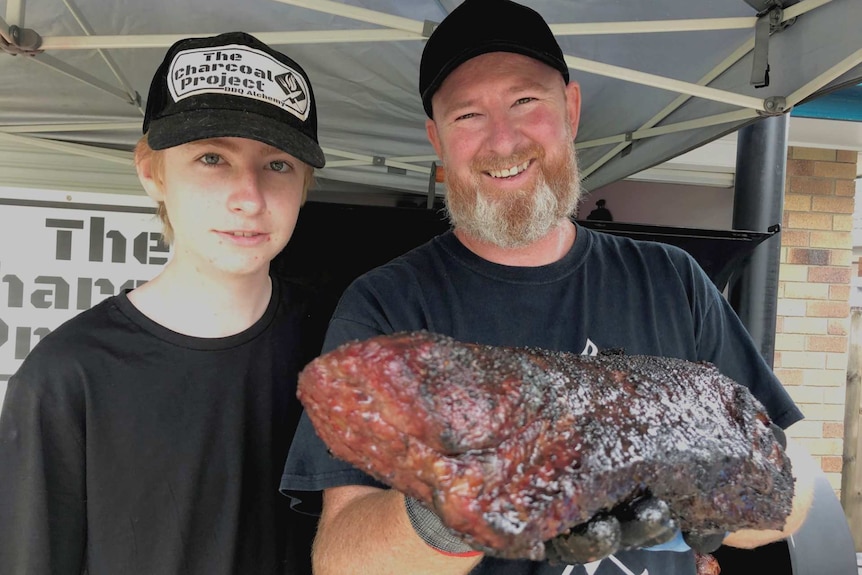 A man stands next to a boy, while holding a giant piece of barbecue-cooked meat